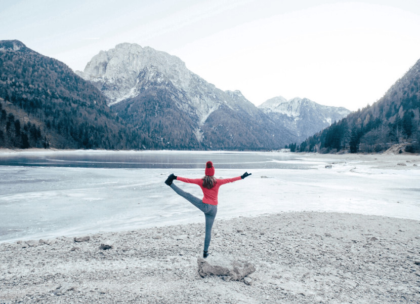 Woman doing a yoga pose in front of an alpine lake (wearing leggings, a pink shirt, gloves, and a beanie)